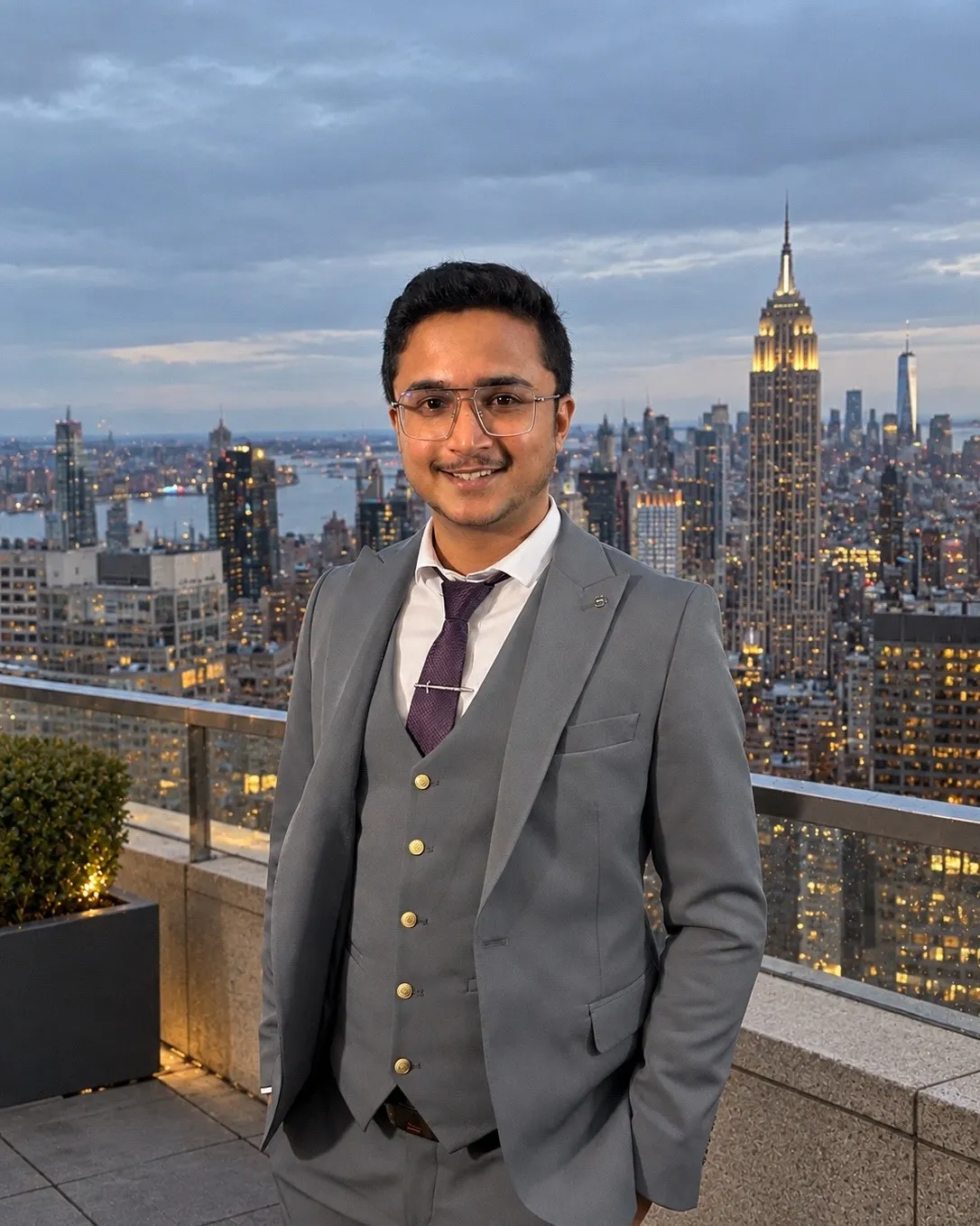 Anuj, the person who builds Solvia Works sites, in a grey suit and glasses on a rooftop, with the New York skyline behind him at dusk